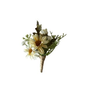 以下が含まれることがあります： クリーム色と黄色の人工花をあしらったブートニア。緑の葉がアクセントになっています。素朴で自然な美しさを生み出すために、紐で巻かれた茎で固定されています。結婚式や特別なイベントに最適です。
