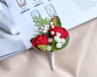 Puede incluir: Un pequeño boutonniere floral rojo y blanco con una cinta de satén blanca. El boutonniere está hecho de flores de seda y es perfecto para una boda u otra ocasión especial.
