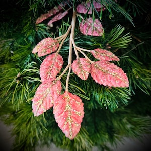 May include: Close-up of a decorative branch with pink leaves and gold glitter accents. The leaves are arranged on a light brown stem, set against a backdrop of green pine needles. This festive piece is suitable for holiday decor.