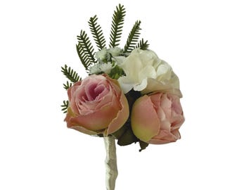 Puede incluir: Un prendido floral con dos rosas color melocotón y una flor blanca, acentuado con ramitas verdes. Los tallos están envueltos en una tela color crema, creando un aspecto clásico y elegante para bodas o eventos especiales.