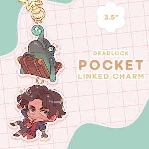 May include: A 3.5 inch acrylic keychain featuring a cartoon character with brown hair and a blue scarf, and a green frog sitting on a brown suitcase. The keychain is labeled "DEADLOCK POCKET LINKED CHARM".