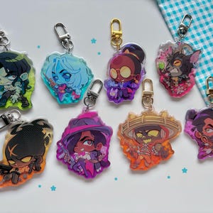 Pode incluir: Uma coleção de oito chaveiros de acrílico com desenhos coloridos de personagens de desenhos animados. Cada chaveiro tem um fecho prateado ou dourado. Os personagens têm várias roupas e expressões, com cores vibrantes como roxo, laranja e azul.