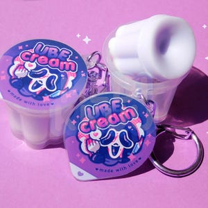 May include: A collection of Ube Cream keychains and a small container with a white, ribbed object inside. The keychains have a purple and blue design with the words "UBE Cream" and the phrase "made with love". The background is a solid pink color.