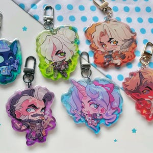 May include: A collection of seven acrylic keychains featuring colorful anime-style character designs. Each keychain has a silver clasp. The characters include a cat, a woman with white hair, a man with a beard, a unicorn, and a devil.