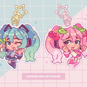 May include: Two colorful anime character keychains with gold star charms. The keychain on the left features a green-haired character holding a green vegetable. The keychain on the right features a pink-haired character with a cherry headband. The text "Hatsune Miku Keychains" is at the bottom of the image.