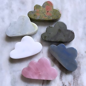 May include: Six cloud-shaped polished stones in various colors, including green, blue, white, pink, and gray, on a white marble surface.