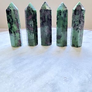 May include: Five green and black gemstone points, also known as green aventurine, are arranged in a row on a white surface.