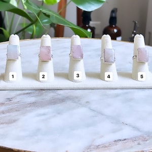 May include: Five silver rings with different colored rectangular stones on display. The stones are pink, light blue, and white. The rings are numbered 1 through 5.
