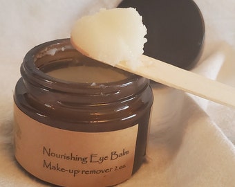 May include: A brown glass jar with a white label that reads "Nourishing Eye Balm Make-up remover 2 oz." A wooden spatula with a scoop of white balm is resting on the jar.