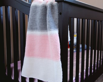 Może przedstawiać: Szary, różowy i biały kocyk dla niemowląt w stylu crochet, drapowany na czarnym łóżeczku.