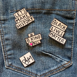 May include: Four pins on a blue denim background. The pins are white with black lettering. The pins say "Let's Make History", "#B Kind All", "Black Lives Matter", and "BLM".