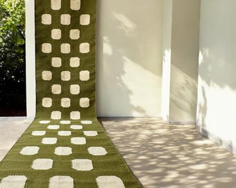 May include: A long, olive green rug with a repeating pattern of large, off-white squares. The rug is hanging from a metal rod and extends across a light-colored floor. The design is symmetrical and modern, suitable for home decor.