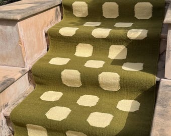 May include: A green and cream-colored stair runner with a repeating pattern of square shapes. The runner is made of a woven material and is installed on a set of outdoor stairs. The color scheme is neutral and would complement many home decor styles.