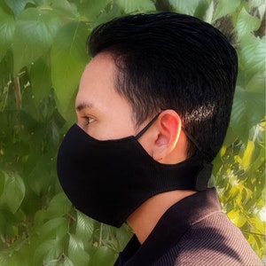 Könnte beinhalten: Eine schwarze Gesichtsmaske mit gerippter Textur, verstellbaren Ohrschlaufen und einem Nackenriemen. Die Maske ist so konzipiert, dass sie Nase und Mund bedeckt und Schutz bietet. Der Hintergrund zeigt grünes Laub.