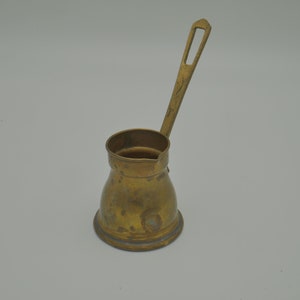 Peut inclure: Une petite cafetière en laiton avec une longue poignée. La cafetière a un corps arrondi et un bord évasé.