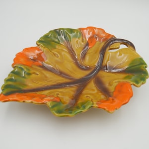 Könnte beinhalten: Eine keramikförmige Blattschale mit Griff und glänzendem Finish. Die Schale hat eine Farbpalette aus Grün, Orange und Gelb mit braunen Details. Das Design ahmt die natürliche Form eines Blattes nach, geeignet für dekorative oder funktionelle Zwecke.