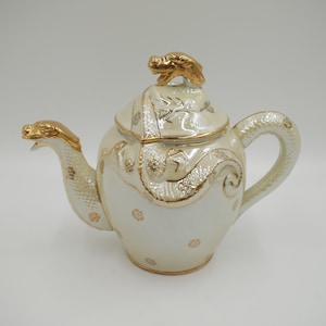 Peut inclure: Une théière en céramique blanche avec des accents dorés et un motif de dragon sur le couvercle et le bec.