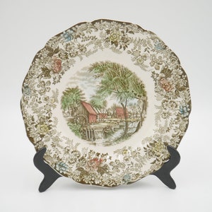 May include: A white ceramic dinner plate with a brown and green floral border and a central image of a red gristmill with a water wheel. The plate is on a black stand.