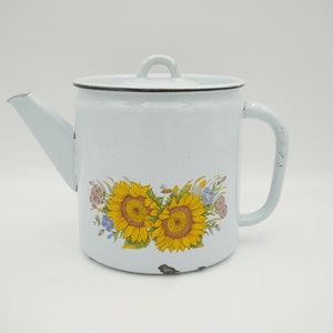 May include: A pale blue enamel teapot with a lid and handle. The teapot is decorated with a floral design featuring two sunflowers and other colourful flowers. It has a spout and is likely vintage.