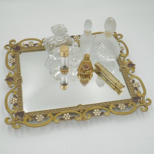 Op de afbeelding: Een antieke toilettafel met een spiegeloppervlak, met verschillende parfumflesjes. De lade heeft een goudkleurig frame met sierlijke details en decoratieve accenten. De parfumflesjes variëren in vorm en grootte, met heldere en matglazen opties.
