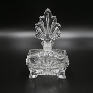 Può includere: Un flacone di profumo trasparente vintage con un tappo a forma di foglia. La bottiglia quadrata ha un design in vetro tagliato e poggia su quattro piedini. Il tappo è decorato con un motivo a ventaglio.