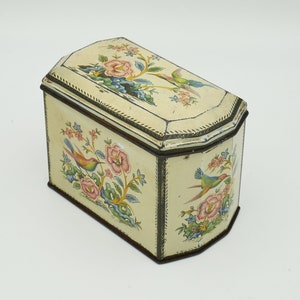 Può includere: Una scatola di latta vintage con sfondo crema e design floreale. La scatola presenta due uccelli colorati e fiori rosa.