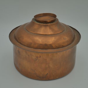 Op de afbeelding: Een koperen pot met een deksel. De pot heeft een rond lichaam en een licht uitlopende rand. Het deksel heeft een kleine knop bovenop.