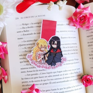 Peut inclure: Gros plan d'un livre avec un marque-page rouge représentant deux personnages d'anime. Le marque-page est placé sur une page avec le texte "El Circo de la Noche" et "Widget" en espagnol. Les personnages sont une fille blonde avec une robe rose et une fille aux cheveux noirs avec une robe noire. Le livre est entouré de roses roses.