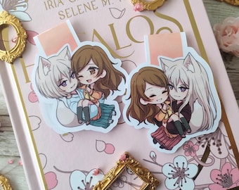 Puede incluir: Dos marcadores de personajes de estilo anime con un lobo blanco y una chica con cabello castaño. Los marcadores están sobre un fondo rosa con un patrón floral. El texto "IRIA G. PARENTE SELENE M. PA" es visible en el fondo.