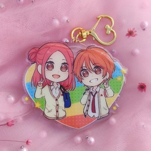 Puede incluir: Un llavero en forma de corazón con un fondo colorido presenta dos personajes de anime. Los personajes son una niña con cabello rosado y un niño con cabello naranja. Ambos llevan uniformes escolares y sonríen.