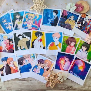 May include: A selection of white polaroid-style prints showcasing vibrant anime and cartoon illustrations. The images feature diverse characters and scenes, with varied styles and themes. The prints are arranged on a wooden surface with decorative embellishments.