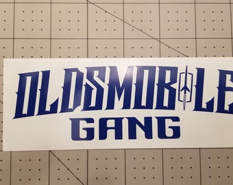 May include: A white sticker with the words "OLDSMOBILE GANG" in blue, stylized lettering. The word "OLDSMOBILE" is arched, and the word "GANG" is below it. A logo of a rocket is between the letters "E" and "L".