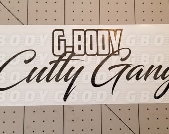 May include: A white sticker with the words "G-BODY" in a bold, blocky font above "Cutty Gang" in a flowing script. The text is black, and the background is white.