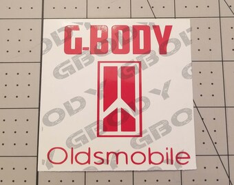 May include: A square white sticker with red text and graphics. The text "G-BODY" is at the top, with "Oldsmobile" at the bottom. A red graphic in the center features a vertical rectangle with a peace sign.