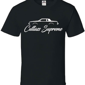 May include: Black t-shirt featuring a white outline of a classic car and the words "Cuttass Supreme" in a cursive font. The shirt has a crew neck and short sleeves. The shirt is made of a soft material.