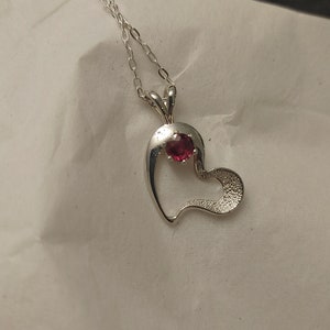 May include: A silver heart-shaped pendant necklace with a small red gemstone in the center.