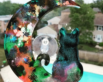 May include: A crescent moon-shaped suncatcher featuring a silhouette of a cat sitting at the base. The piece is crafted from resin with a swirling blend of black, green, orange, and purple colors, accented with small black spider shapes and glitter.