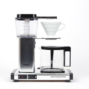 May include: A silver and black Moccamaster coffee maker with a clear water reservoir and a glass carafe. A white ceramic pour-over cone sits above the carafe. The machine has a power switch and a warming plate.