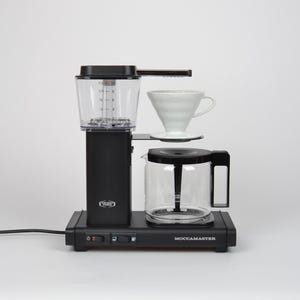 May include: A black Technivorm Moccamaster coffee maker featuring a clear water reservoir with measurement markings, a white ceramic cone dripper, and a glass carafe with a black handle. The base includes two black power switches and the brand name.