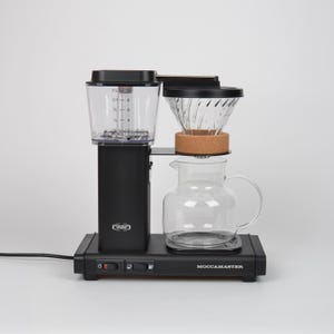 May include: A matte black Technivorm Moccamaster coffee maker featuring a clear water reservoir with markings up to 10 cups, a glass filter cone with a cork collar, and a glass carafe. The base includes a power switch and a heating element control.