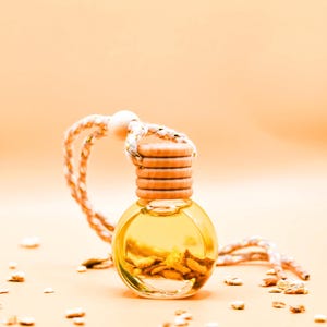 Op de afbeelding: Een kleine, heldere glazen fles gevuld met een gele vloeistof en gedroogde botanische producten. De fles heeft een houten dop en een gevlochten touw om op te hangen. De achtergrond is perzikkleurig, met kleine stukjes gedroogde botanische producten.