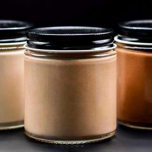 Könnte beinhalten: Drei Gläser mit schwarzen Deckeln und weißen Rändern, gefüllt mit brauner Creme in verschiedenen Farbtönen. Die Gläser sind in einer Reihe vor einem dunklen Hintergrund angeordnet und zeigen die verschiedenen Farben des Produkts.