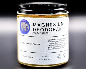 Könnte beinhalten: Ein Glasbehälter mit Magnesium-Deodorant, schwarzem Deckel und weißem Etikett. Das Etikett zeigt ein blaues kreisförmiges Logo mit einem geometrischen Elefanten, den Text Magnesium Deodorant Cozy Nights, Patchouli Myrrh Ginger und 113g.