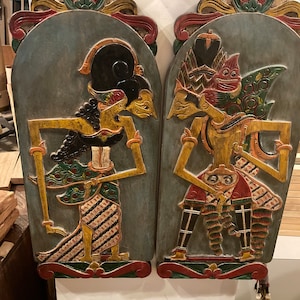 May include: Two intricately carved wooden panels depicting traditional Indonesian shadow puppets. The panels are painted in vibrant colors, including red, green, yellow, and blue. The figures are depicted in a stylized manner, with exaggerated features and flowing garments.