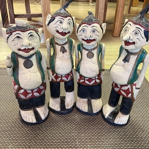 May include: Four wooden figurines of smiling, whimsical characters with large ears, painted in white, red, green, and black. The figurines are standing on a brown patterned rug.