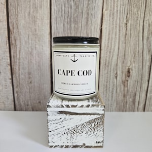 Op de afbeelding: Een glazen potkaars met een zwart deksel, met het opschrift "CAPE COD" met de geur "Citrus Oakmoss Violet". De kaars staat op een klein, wit, verweerd houten blok. De achtergrond is een lichtbruine houten muur.