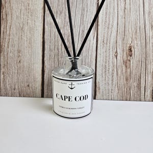Puede incluir: Botella difusora de vidrio transparente con varillas negras. La etiqueta dice "CAPE COD" con un símbolo de ancla y "Citrus Oakmoss Violet". La botella está sobre una superficie blanca con fondo de madera.