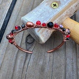 May include: A copper wire cuff bracelet featuring a series of beads in red, black, and metallic bronze tones. The wire is intricately wrapped around the beads, creating a textured band with a circular opening. The design incorporates faceted crystals.