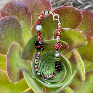 May include: A red, black, gold, and silver beaded bracelet with flower-shaped charms. The bracelet is on a green succulent plant.