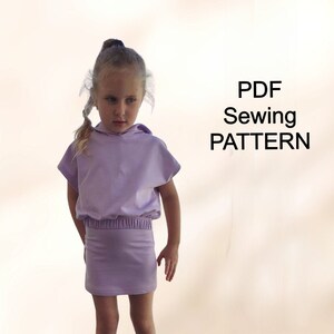 May include: A PDF sewing pattern for a lilac hooded top and skirt set for a child. The set features a short-sleeved top with a hood and a skirt with an elastic waistband.